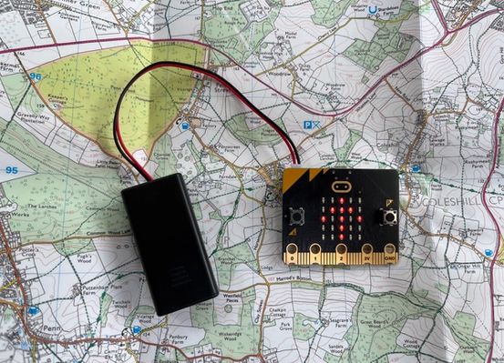 a micro:bit and attached battery pack resting on top of a map with an up arrow displayed on the micro:bit LED display